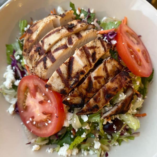 Grilled chicken salad with tomatoes, feta cheese, and olives in a white bowl at Fora Cafe.