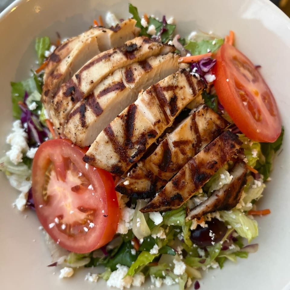 Grilled chicken salad with tomatoes, feta cheese, and olives in a white bowl at Fora Cafe.