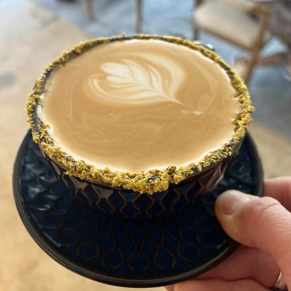 A hand holds a dark blue Fora Cafe latte with intricate latte art and a pistachio-dusted rim.